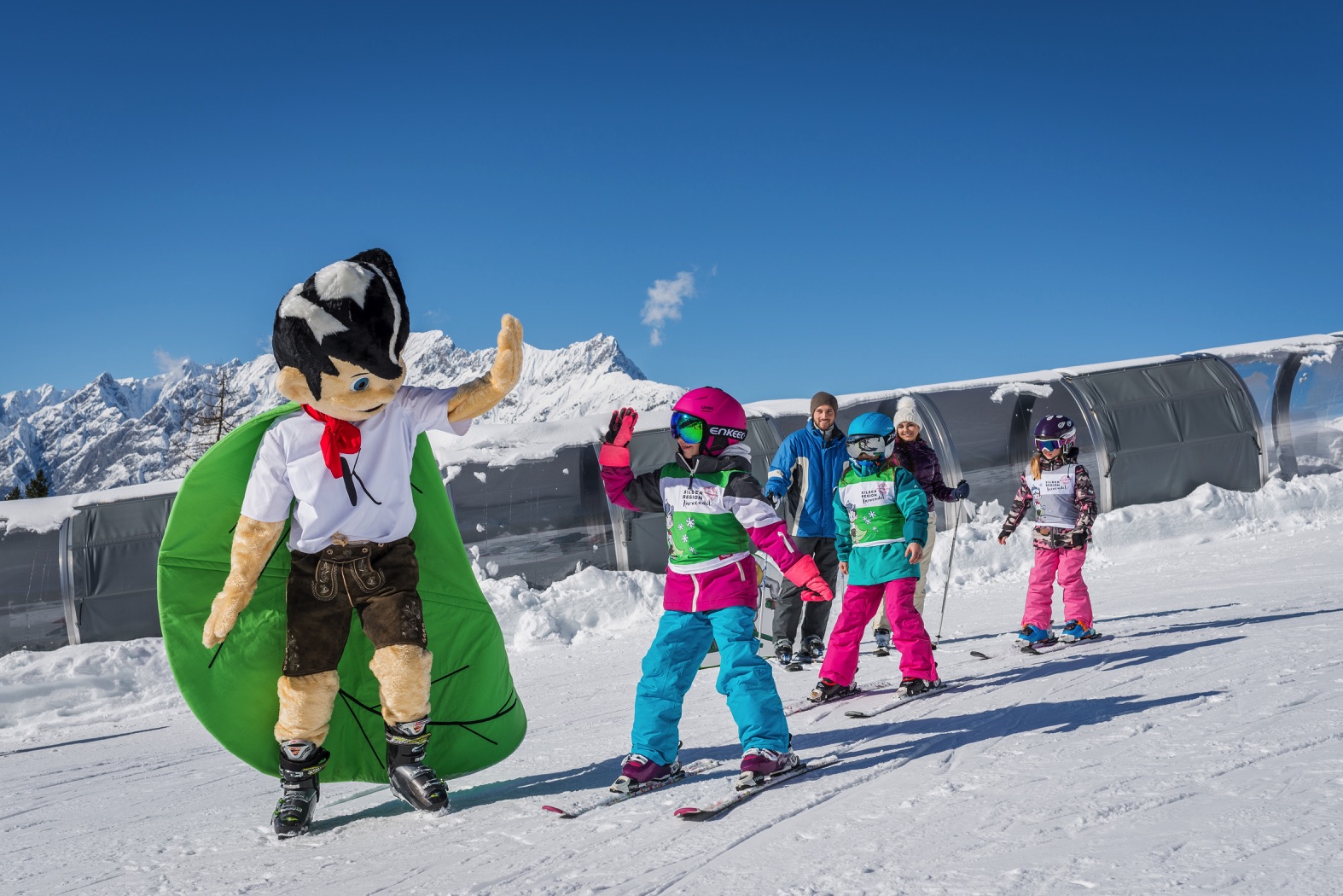 Kinder auf Schiern mit dem Maskottchen der Silberregion Karwendel vor verschneiten Bergen